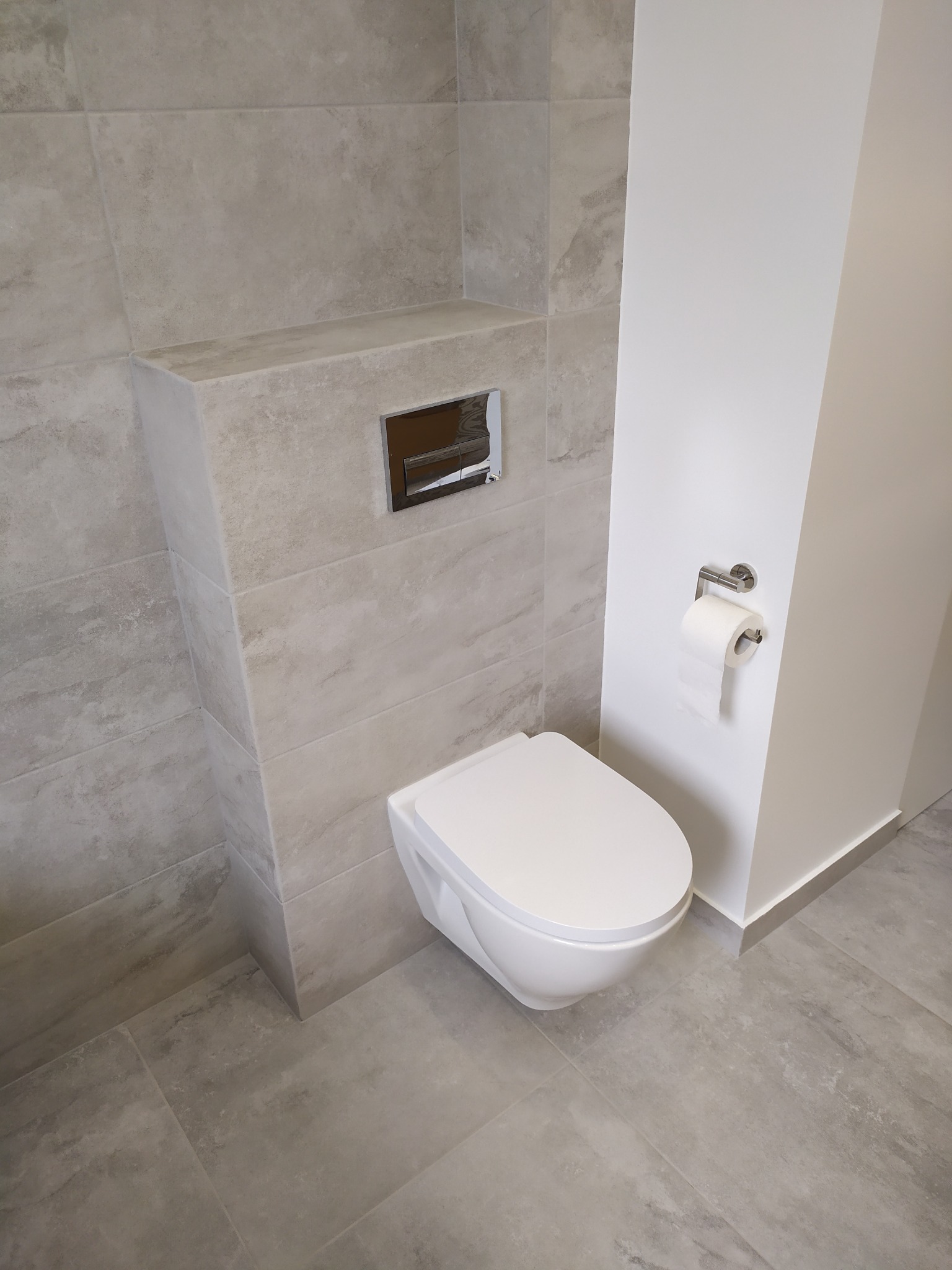 Nowoczesna łazienka w odcieniach szarości z podwieszaną miską WC i chromowanym przyciskiem spłukującym oraz uchwytem na papier toaletowy. Płytki imitujące kamień na ścianach i podłodze.