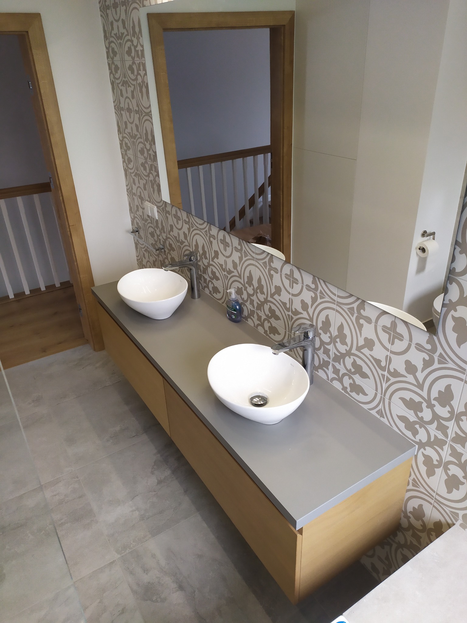 Nowoczesna łazienka we Wrocławiu z dwoma umywalkami nablatowymi o okrągłym kształcie, szarą szafką pod umywalki z drewnianymi frontami, lustrem z dekoracyjnymi płytkami w tle i szarą podłogą...