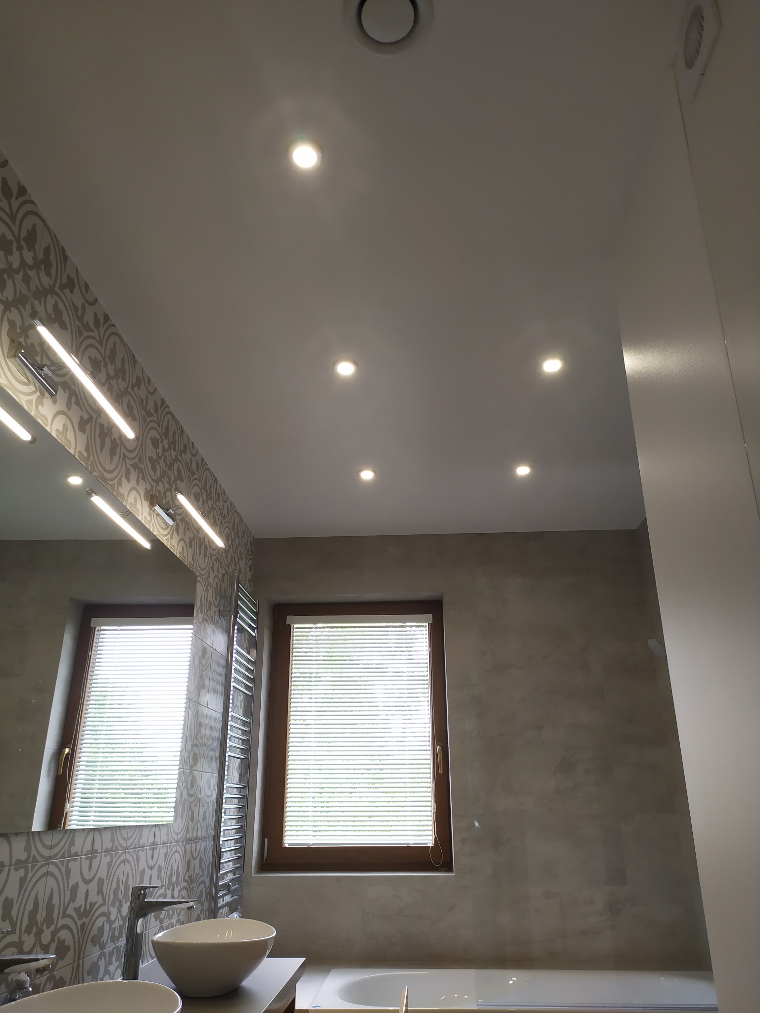 Nowoczesna łazienka z dwoma umywalkami nablatowymi, dużym lustrem z podświetleniem LED, grzejnikiem drabinkowym i oknami z żaluzjami, oświetlona punktowymi lampami sufitowymi