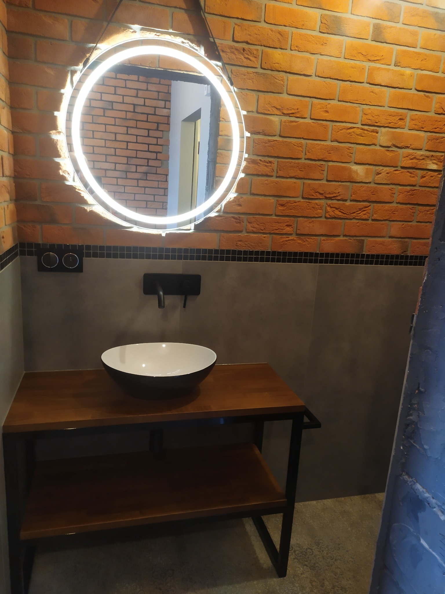 Nowoczesna łazienka w stylu loft: umywalka nablatowa na drewnianym blacie z czarnymi nogami, okrągłe lustro z podświetleniem LED na tle ściany z czerwonej cegły i szarych płytek.