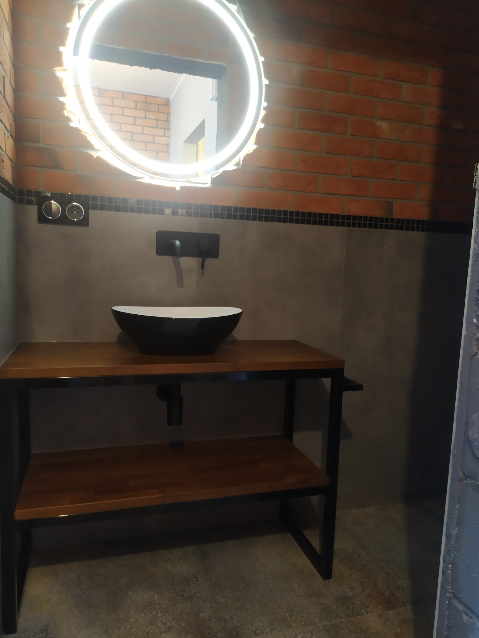 Nowoczesna łazienka z umywalką w kształcie misy na drewnianym blacie z metalową ramą, okrągłe lustro z podświetleniem LED, ściana z czerwonej cegły i szarych płytek.