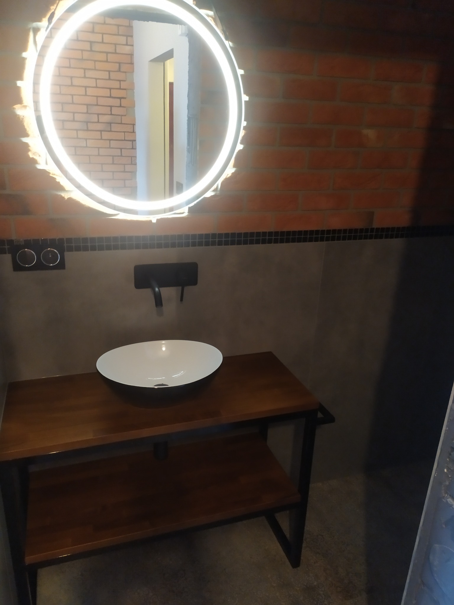 Nowoczesna łazienka z umywalką nablatową na drewnianym blacie z półką, okrągłym lustrem z podświetleniem LED i ścianą z czerwonej cegły.