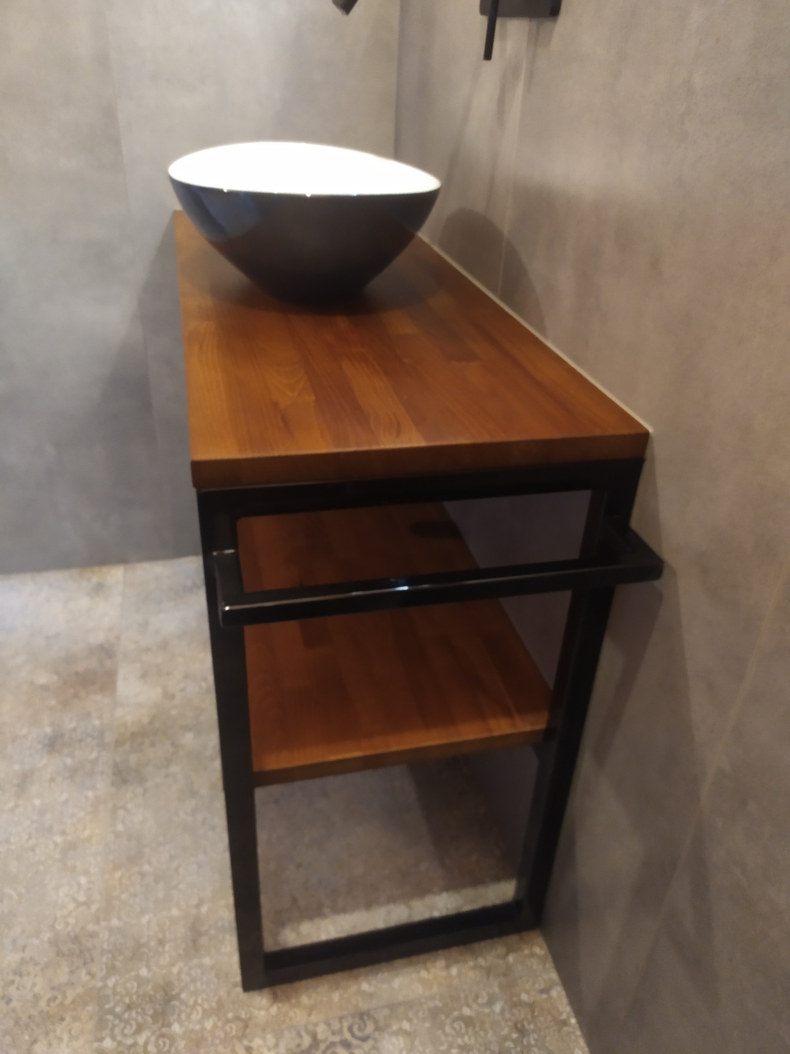 Umywalka nablatowa w kolorze czarno-białym na drewnianym blacie z czarną metalową konstrukcją, widok z góry, szare płytki na ścianie i podłodze.