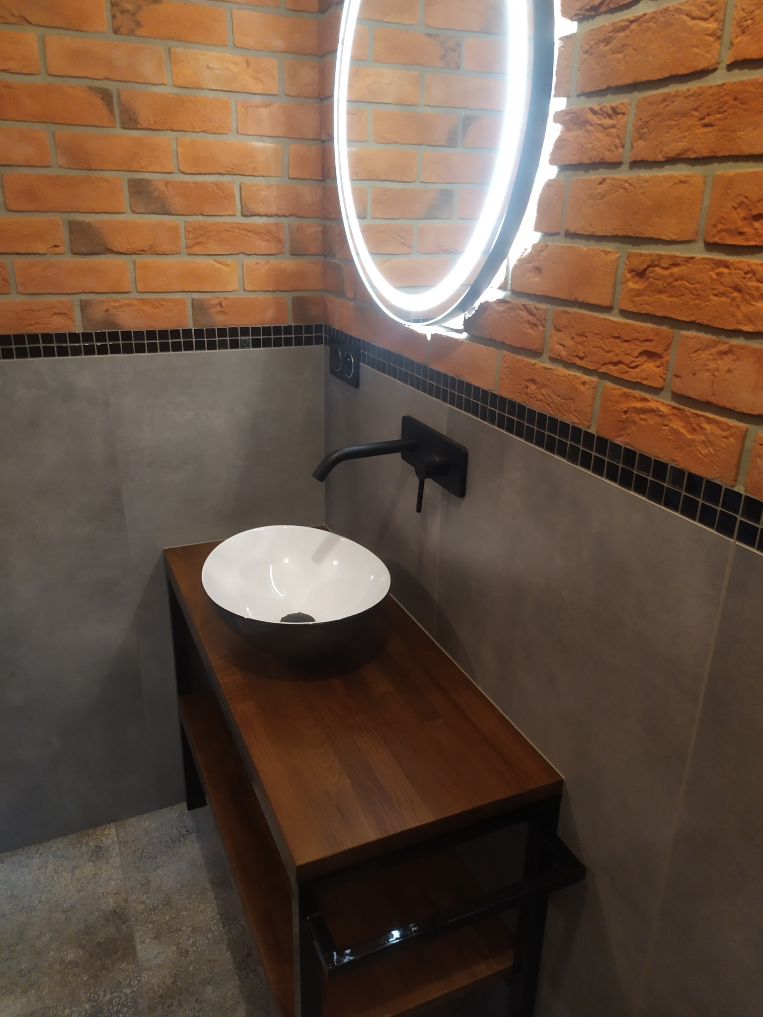 Nowoczesna łazienka z umywalką nablatową, czarną armaturą i podświetlanym lustrem na tle ceglanej ściany.