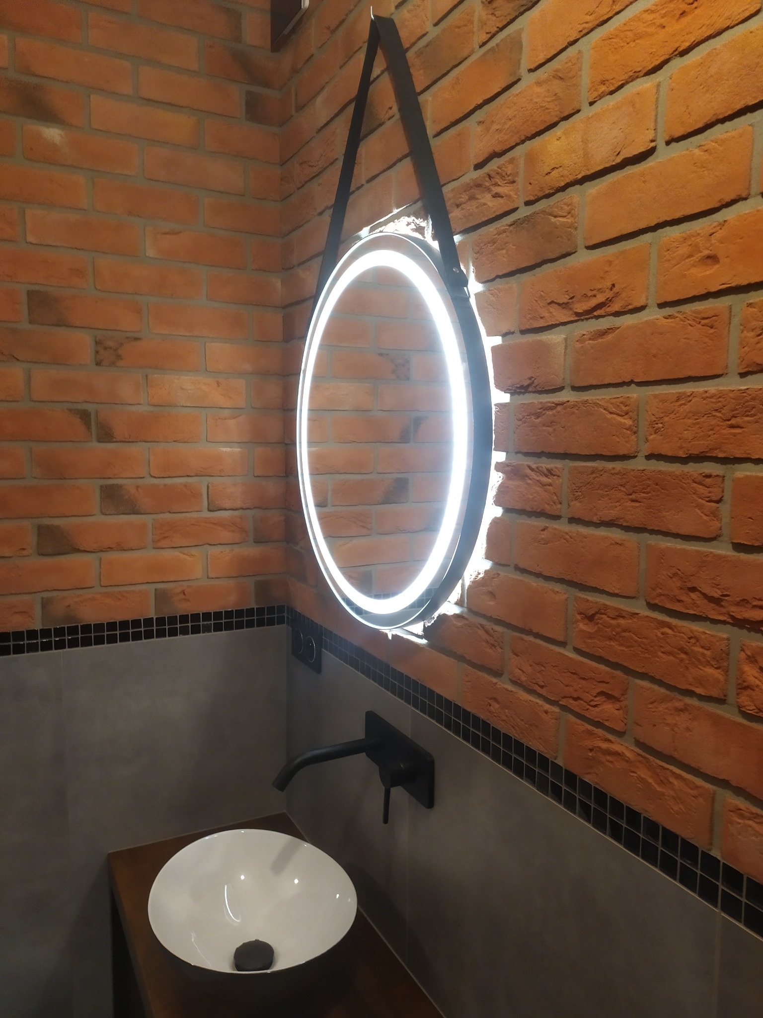 Okrągłe lustro z podświetleniem LED zawieszone na czarnym pasku, zamontowane na ścianie z cegły w łazience z umywalką nablatową i czarną armaturą.