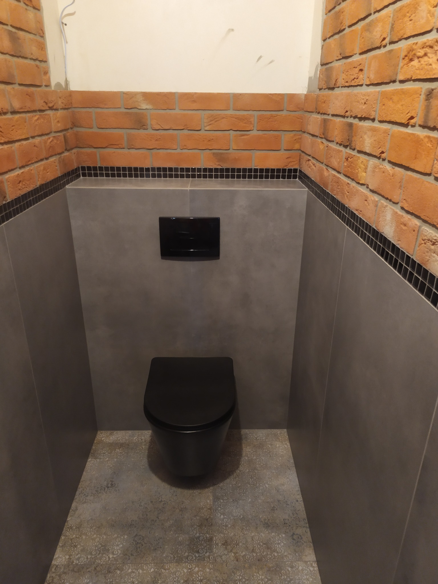 Nowoczesna toaleta w wykończonej łazience, z czarną miską ustępową i przyciskiem spłukującym na tle szarej ściany, obramowana cegłą i czarną mozaiką.