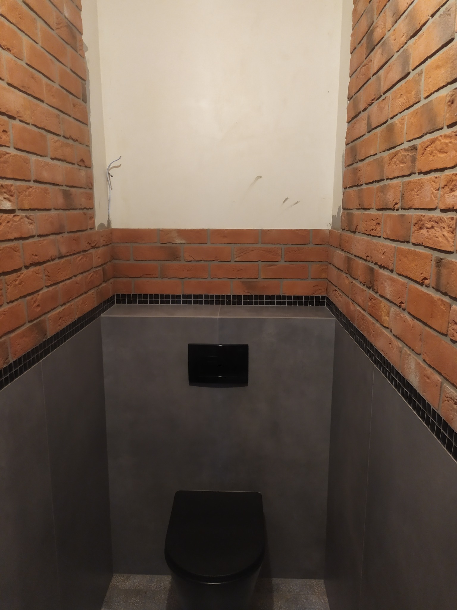Wykończona toaleta w stylu loft z ceglanymi ścianami, szarą zabudową i czarną armaturą, widoczne wystające kable elektryczne.