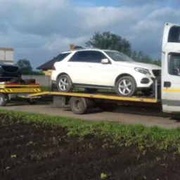 Biała ciężarówka z naczepą transportuje dwa samochody: czarny SUV na żółtej przyczepie i biały SUV na platformie ciężarówki.