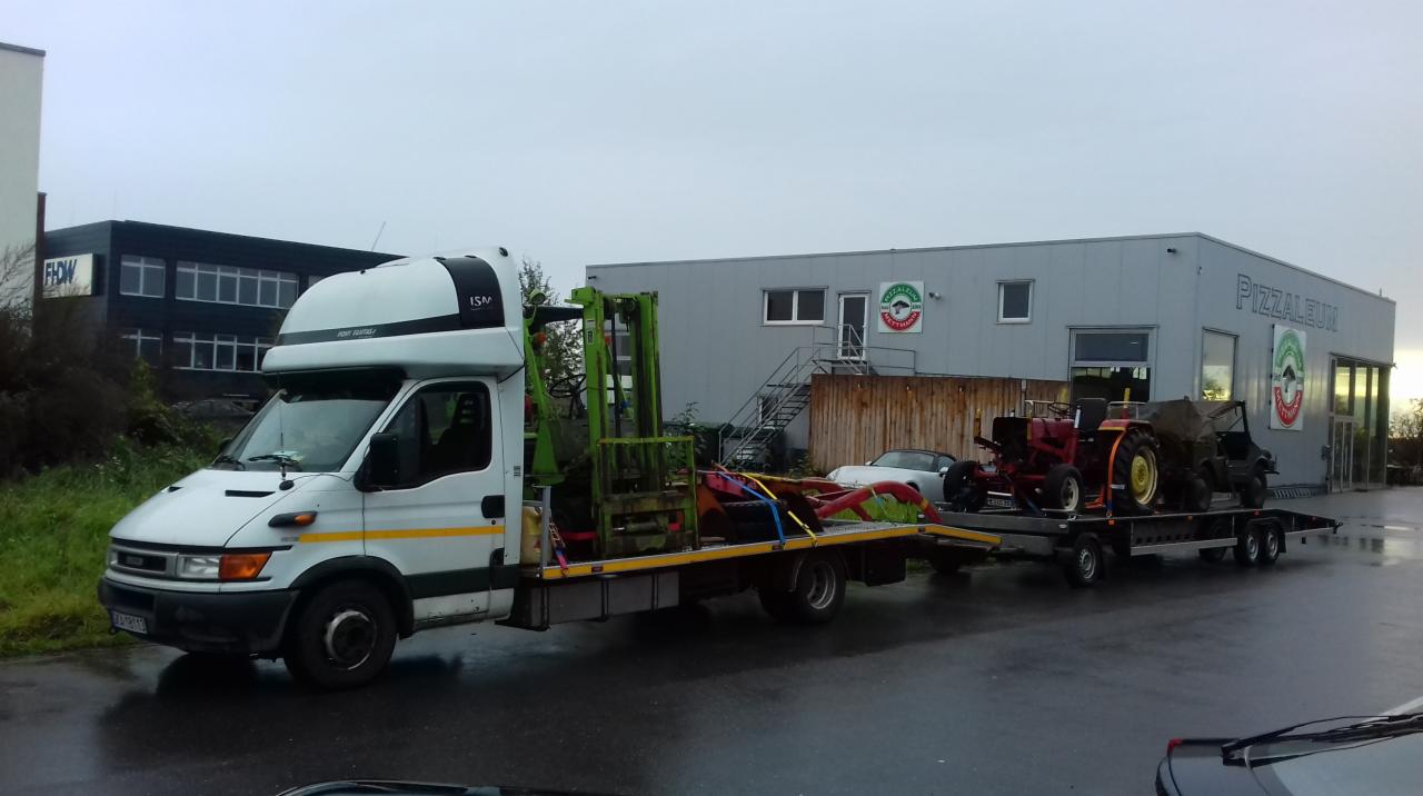 Biała laweta z platformą przewożąca wózek widłowy i na przyczepie dwa traktory na tle budynku z napisem PIZZALEUM w pochmurny dzień.