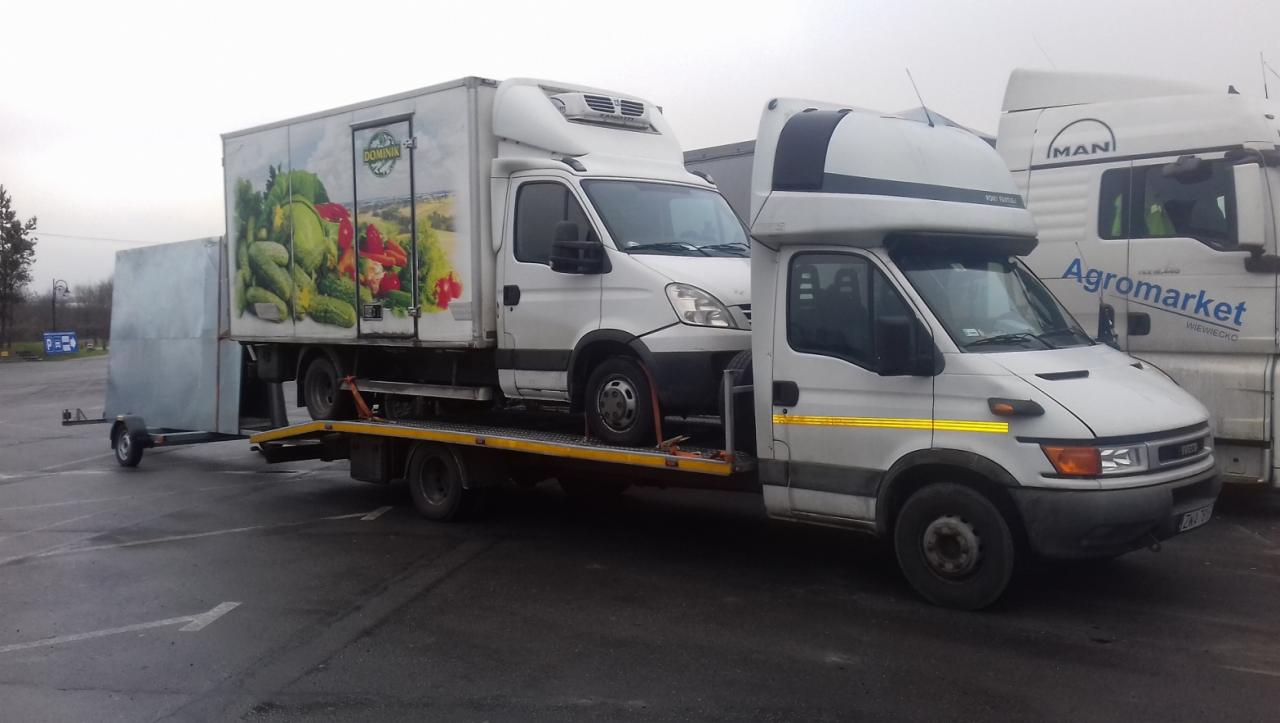 Dwa białe samochody dostawcze Iveco, jeden z nadrukiem warzyw, przewożone na żółtej platformie lawety, z szarym przyczepką z tyłu, na parkingu z widocznym w tle samochodem ciężarowym MAN Agromarket.