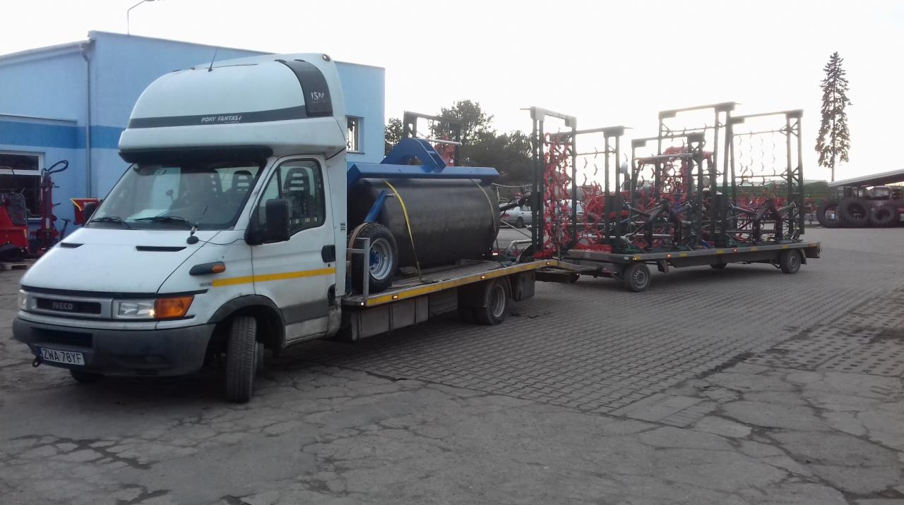 Biały samochód dostawczy z naczepą przewozi walec rolniczy i brony talerzowe na betonowym placu.