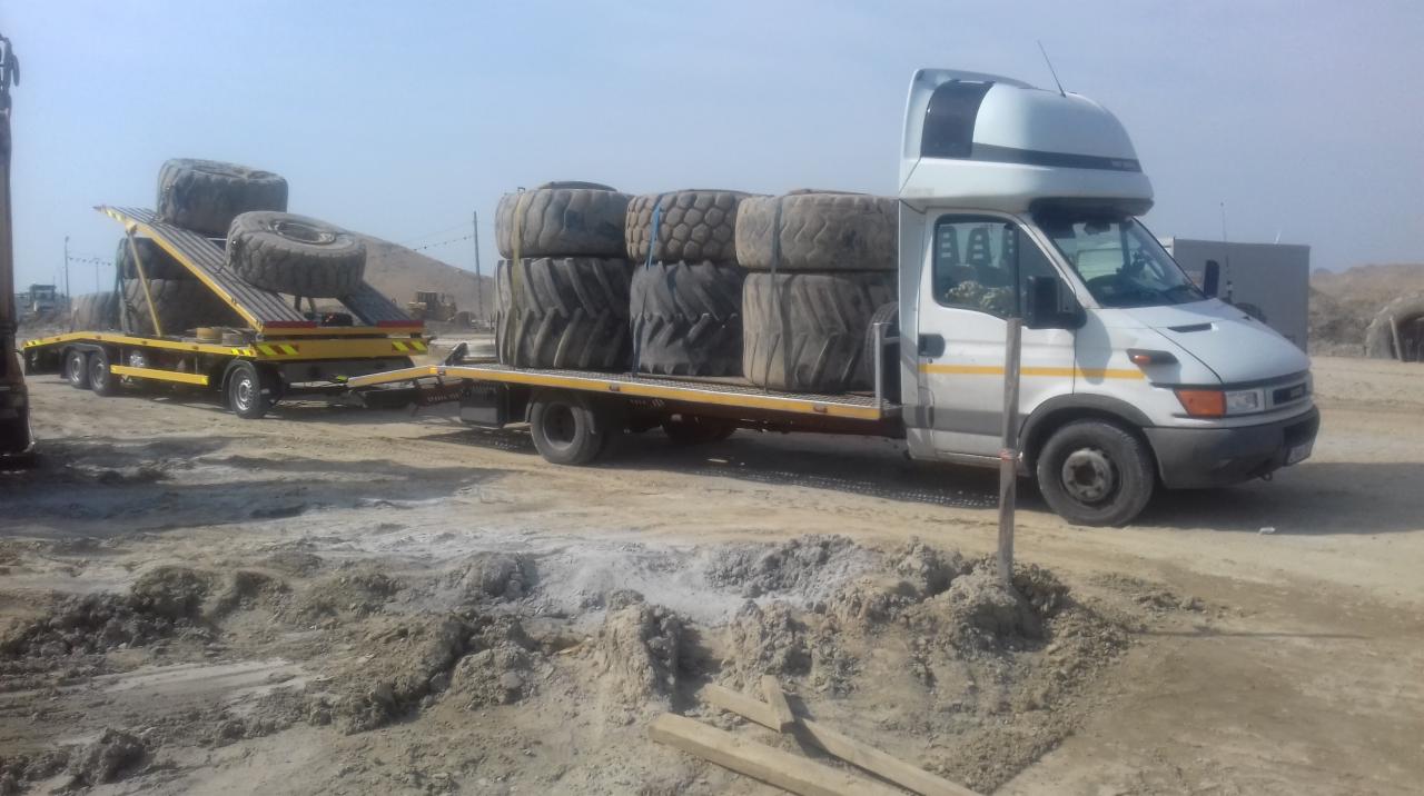 Biała ciężarówka Iveco z platformą transportową i żółtą przyczepą przewozi używane opony rolnicze na placu budowy.