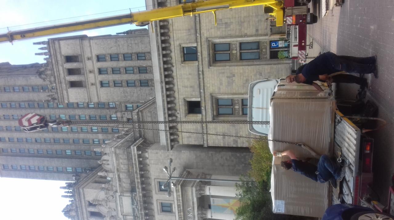 Żółty dźwig podnosi zapakowany ładunek na platformę ciężarówki na tle Pałacu Kultury i Nauki w Warszawie.