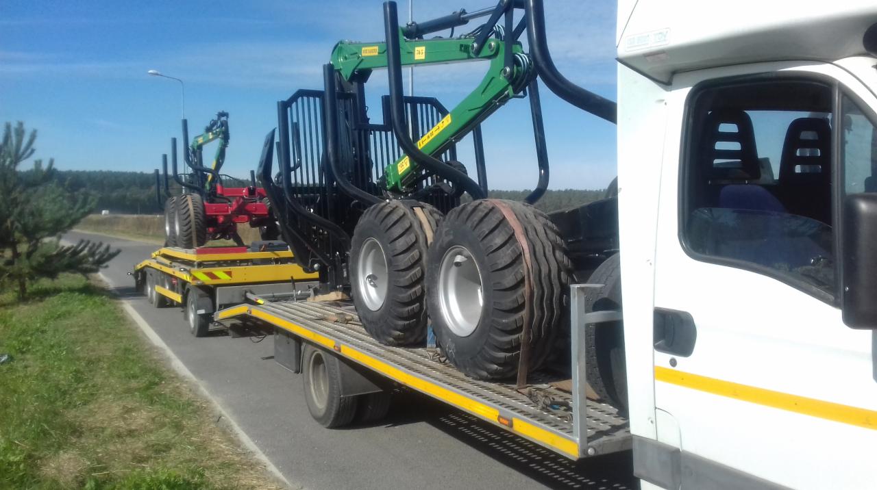 Żółta laweta przewozi dwa duże, zielono-czarno-czerwone ciągniki leśne z chwytakami, widok z boku na tle zieleni i nieba.