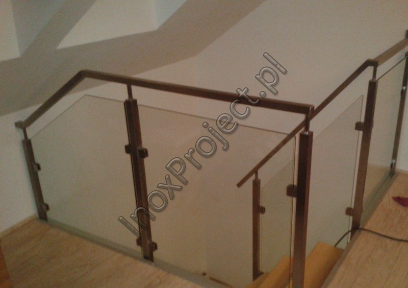 Szklana balustrada ze stalowymi słupkami i poręczą o kwadratowym przekroju, zamocowana na schodach. Widoczne mocowania szkła do słupków.