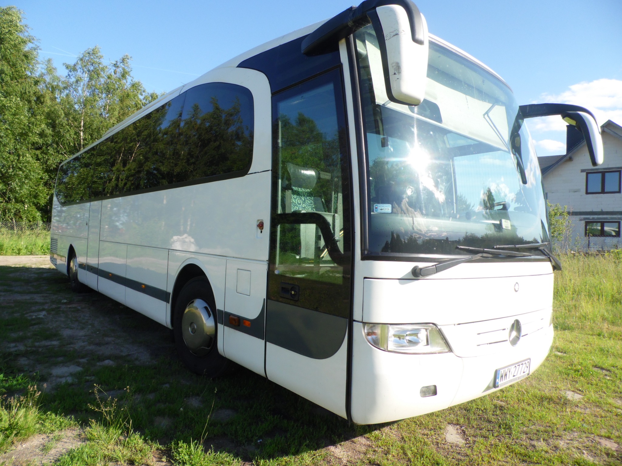 Biały autokar marki Mercedes-Benz z otwartymi drzwiami, zaparkowany na trawie w słoneczny dzień, z widocznym numerem rejestracyjnym WWY 27729.
