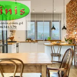 Wnętrze kuchni z drewnianym stołem i krzesłami, ceglaną ścianą oraz oknem z żaluzjami poziomymi; widoczne logo firmy nałożone na obraz.