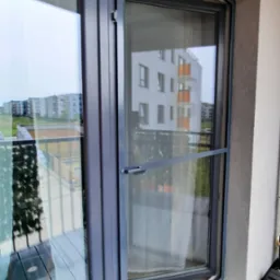 Szara moskitiera ramkowa zamontowana w oknie balkonowym, widok z balkonu na osiedle mieszkaniowe.