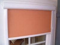 Pomarańczowa roleta materiałowa zwinięta częściowo w oknie z białymi ramami, widoczne zaokrąglone zakończenie kasety rolety.