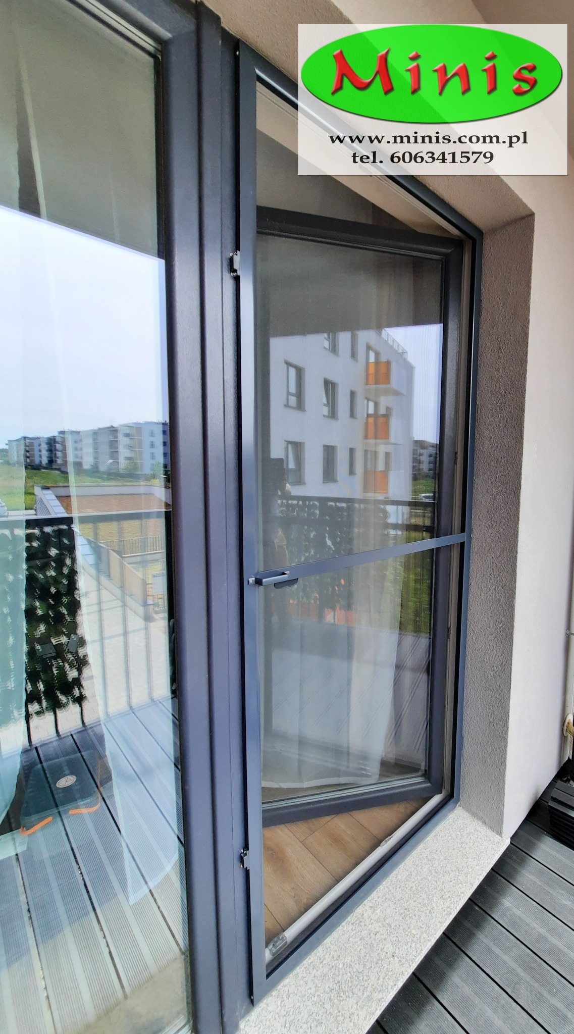 Szara moskitiera ramkowa zamontowana w oknie balkonowym, widok z balkonu na osiedle mieszkaniowe.