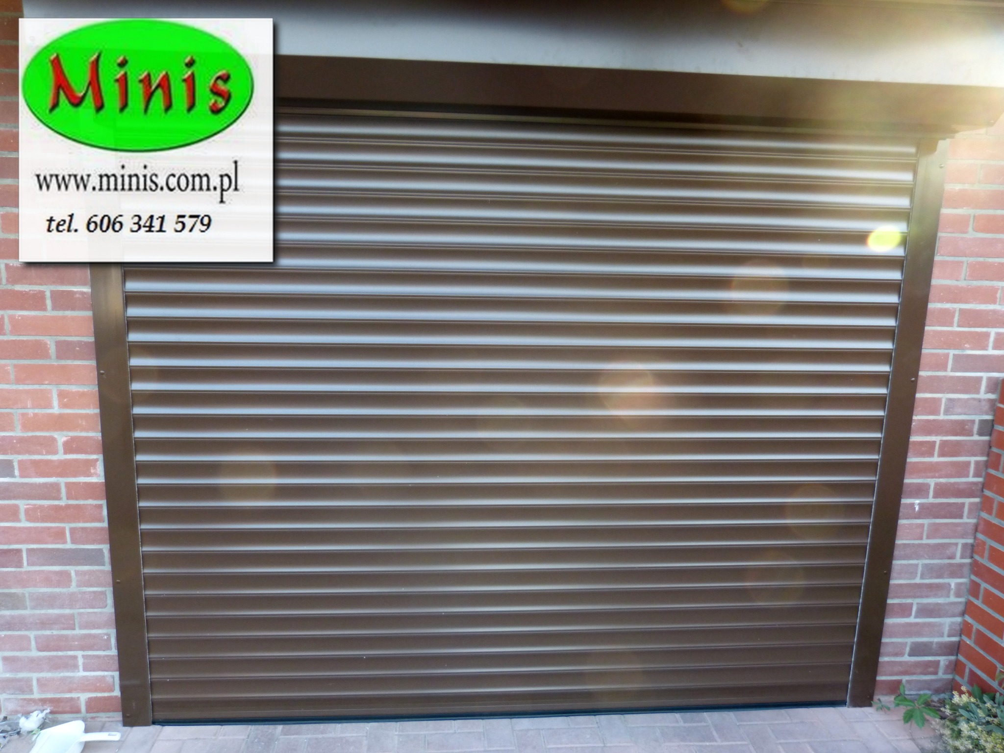 Zewnętrzna, brązowa osłona okienna zamontowana na ceglanym budynku, z widocznym logo firmy i danymi kontaktowymi w lewym górnym rogu.