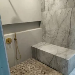 Nowoczesna kabina prysznicowa z marmurową ławką, złotą armaturą i heksagonalną mozaiką podłogową.