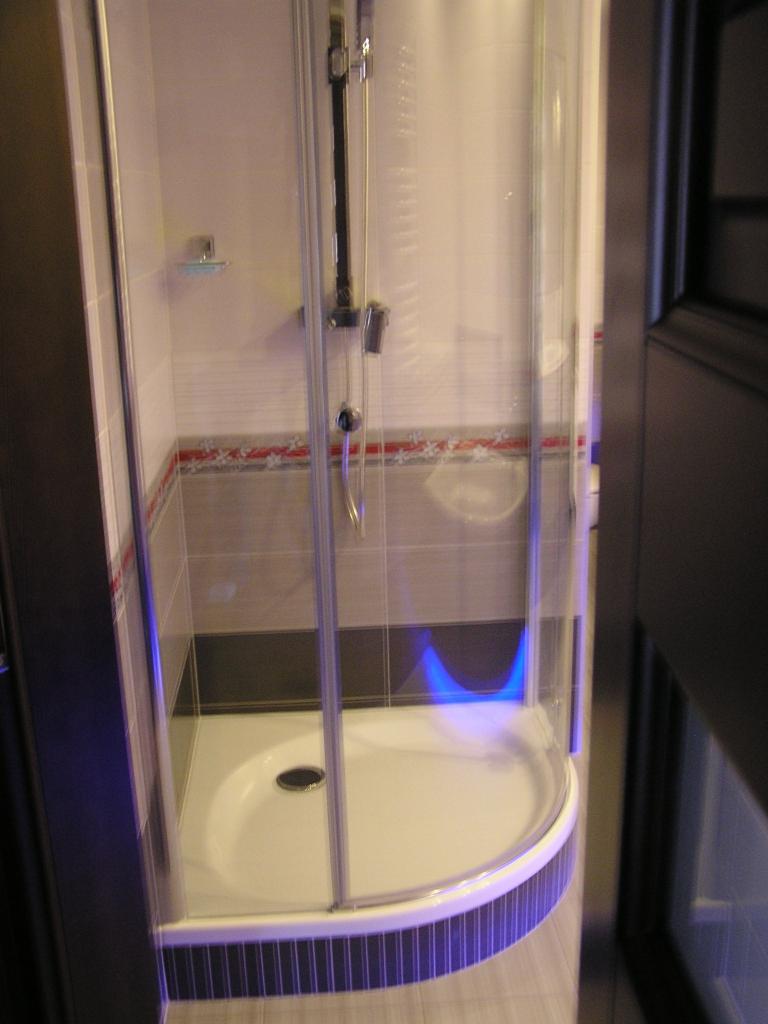 Nowoczesna kabina prysznicowa z białą brodzikiem, szklanymi drzwiami i chromowaną armaturą. Widoczne płytki w odcieniach szarości z dekoracyjnym pasem. Oświetlenie akcentujące.