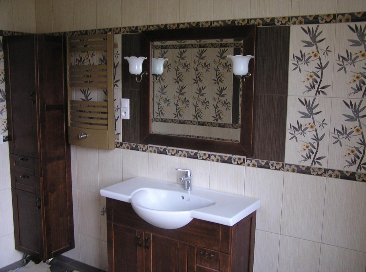 Wykończona łazienka z umywalką w szafce z ciemnego drewna, lustrem w ramie i szafką wiszącą. Na ścianach kremowe płytki z motywem roślinnym i dekoracyjnym pasem.