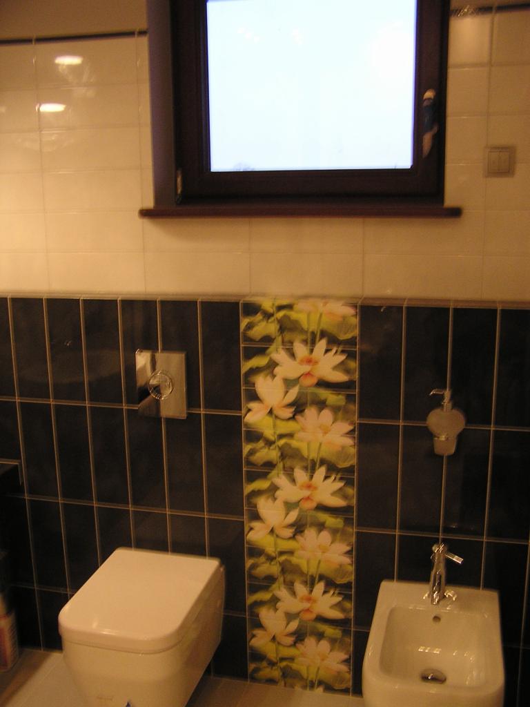 Wykończona łazienka z toaletą i bidetem. Czarne płytki ułożone pionowo, przełamane pasem płytek z motywem kwiatowym. Okno w ciemnej ramie nad płytkami.