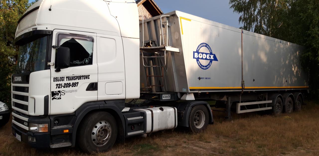 Biała ciężarówka Scania z naczepą Bodex, z widocznym logo firmy transportowej 'Skorpion' i numerem telefonu, zaparkowana na trawiastym terenie.