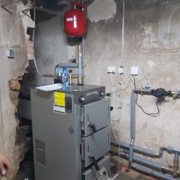 IEMT KOTŁY CO , KLIMATYZACJĘ , POMPY CIEPŁA - Szary piec CO marki DEFRO z czerwonym zbiornikiem wyrównawczym w kotłowni. Widoczne rury instalacji, manometr i butelka wody. Ściany z widocznymi śladami użytkowania.