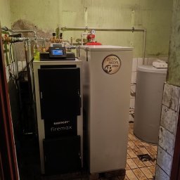 Kocioł Rakoczy Firemax z zbiornikiem buforowym w pomieszczeniu z widocznymi rurami i instalacją. Na kotle naklejka 'Złoty Medal 2024'.