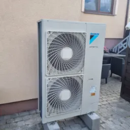 Zewnętrzna jednostka pompy ciepła Daikin Altherma, zamontowana na betonowych podstawach przy ścianie budynku w Iwcu, na tle kostki brukowej i elementu ogrodzenia.