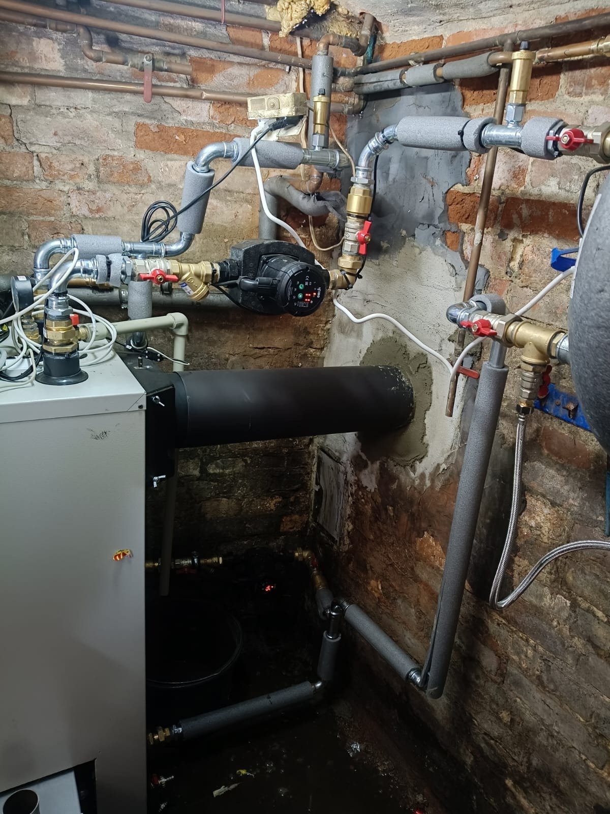 Złożona instalacja grzewcza w piwnicy z ceglanymi ścianami. Widoczne rury, zawory, pompa obiegowa i piec. Część rur zaizolowana termicznie.