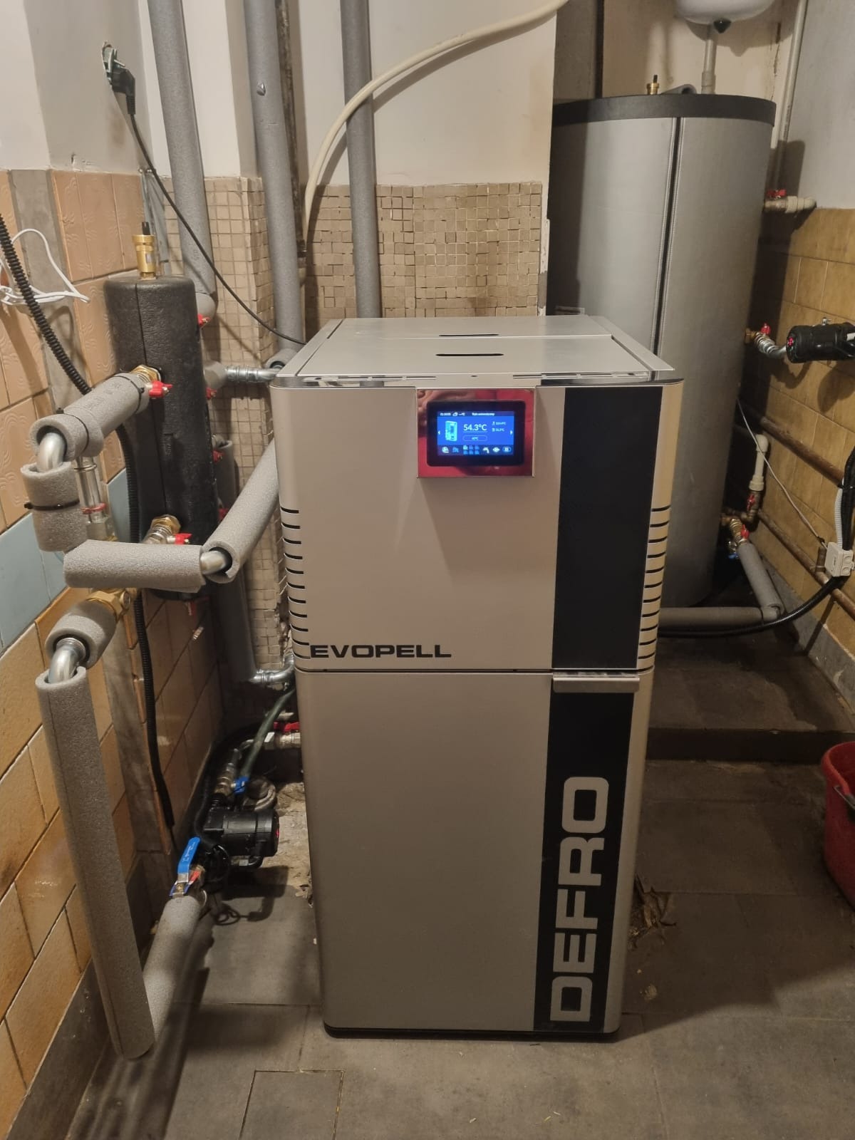 Szary piec Defro Evopell z ekranem dotykowym, widoczne rury instalacji grzewczej i zasobnik wody. Ujęcie w kotłowni z mozaikową okładziną ścienną.