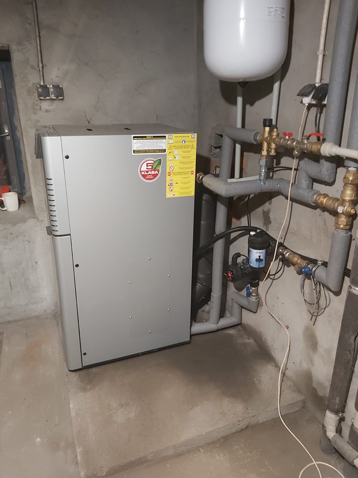 Szary piec CO marki Klaba w piwnicy, z widocznymi rurami i zbiornikiem wyrównawczym. Ściany surowe, betonowe. Instalacja po montażu, gotowa do użytku.