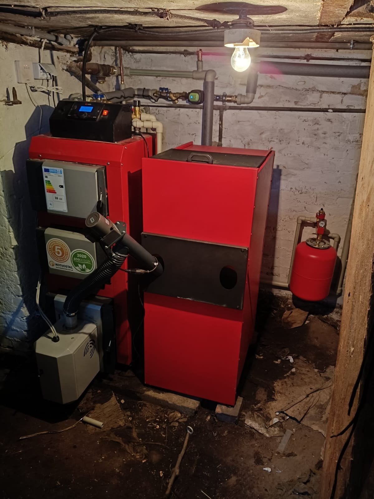 Czerwony piec CO Kotłostal z podajnikiem, zainstalowany w piwnicy z widocznymi rurami i oświetleniem żarówką. Widoczny zbiornik wyrównawczy i elementy instalacji.