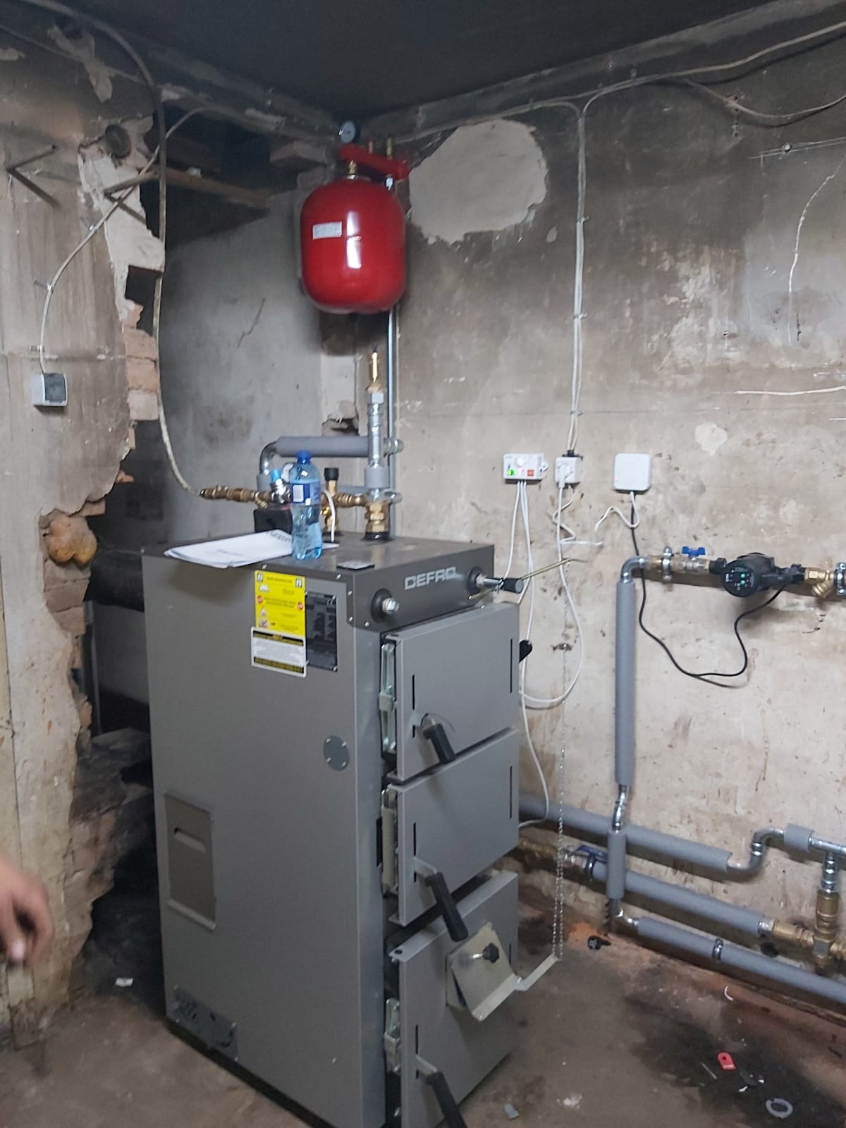 Szary piec CO marki DEFRO z czerwonym zbiornikiem wyrównawczym w kotłowni. Widoczne rury instalacji, manometr i butelka wody. Ściany z widocznymi śladami użytkowania.