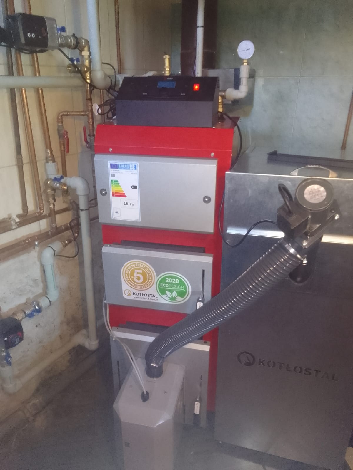 Czerwono-szary piec CO marki Kotłostal z widocznymi naklejkami ECODESIGN i informacją o klasie energetycznej 16kW, podłączony do instalacji w kotłowni.