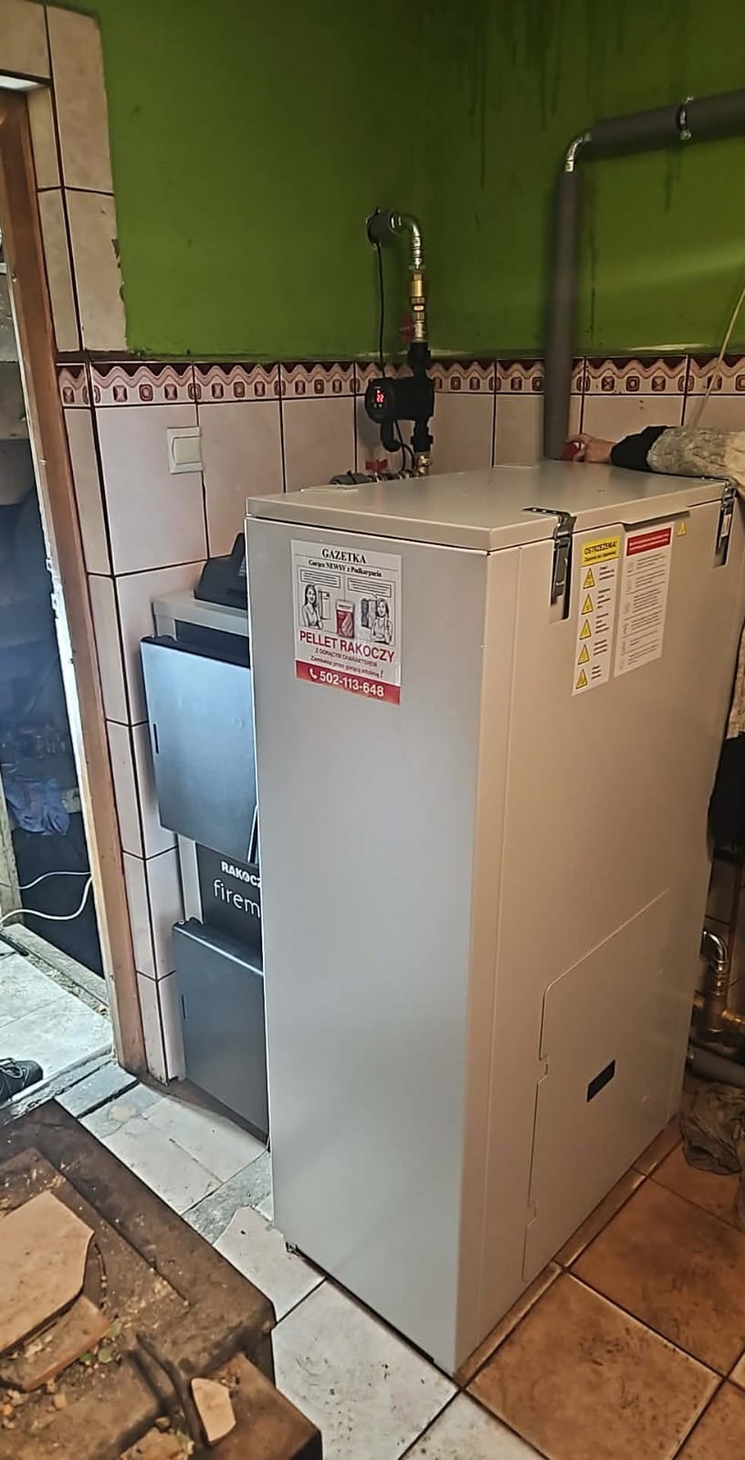 Nowoczesny piec na pellet Rakoczy z widoczną naklejką informacyjną i numerem telefonu, zainstalowany w pomieszczeniu z zieloną ścianą i brązowymi płytkami.