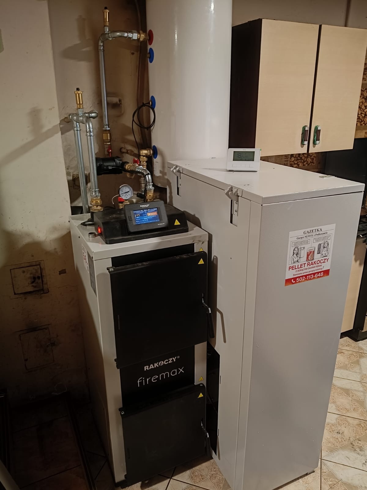 Nowoczesny piec na pellet Rakoczy Firemax z widocznymi rurami instalacji, manometrem i sterownikiem, ustawiony w kotłowni z beżowymi szafkami w tle.