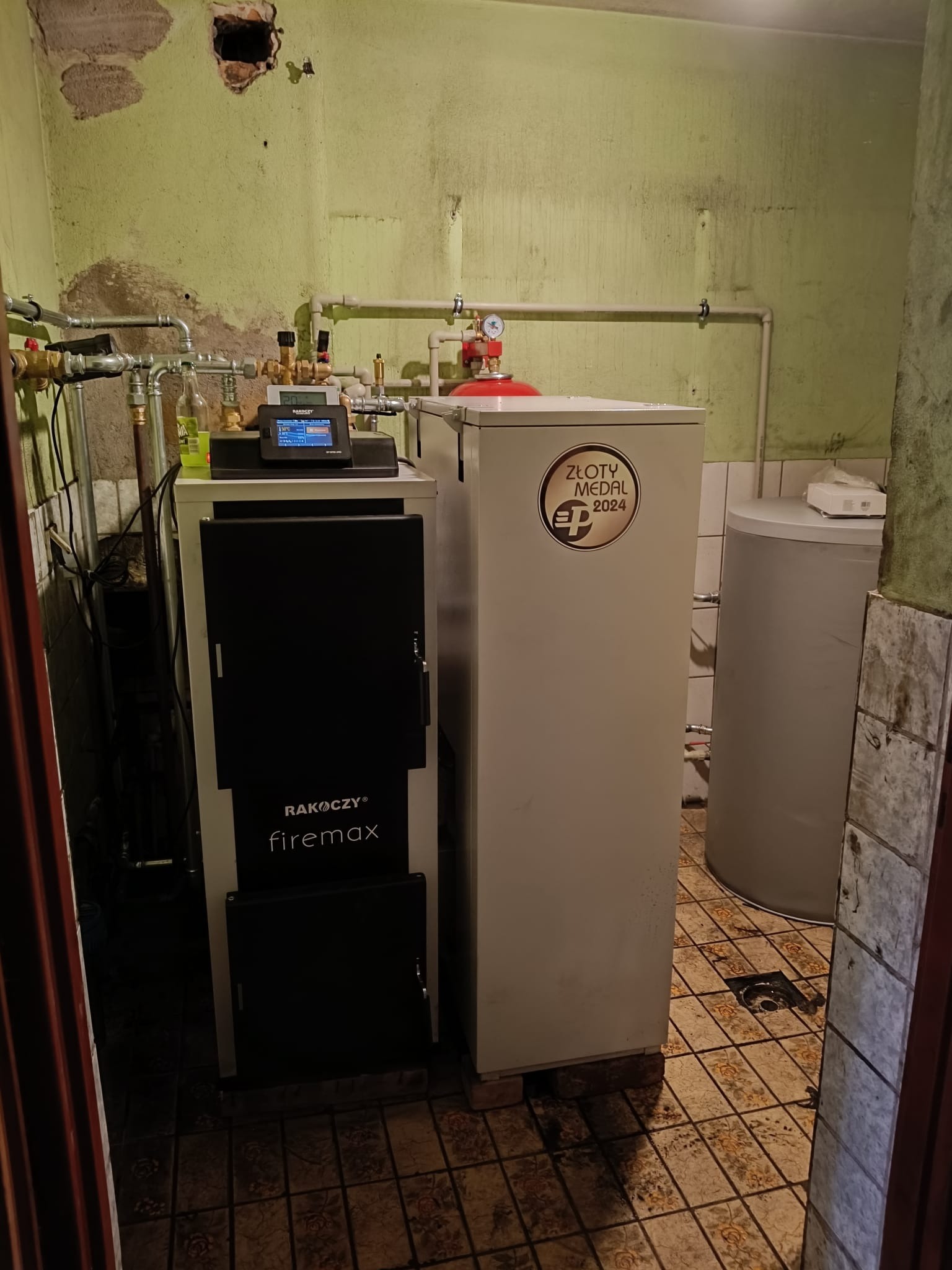 Kocioł Rakoczy Firemax z zbiornikiem buforowym w pomieszczeniu z widocznymi rurami i instalacją. Na kotle naklejka 'Złoty Medal 2024'.