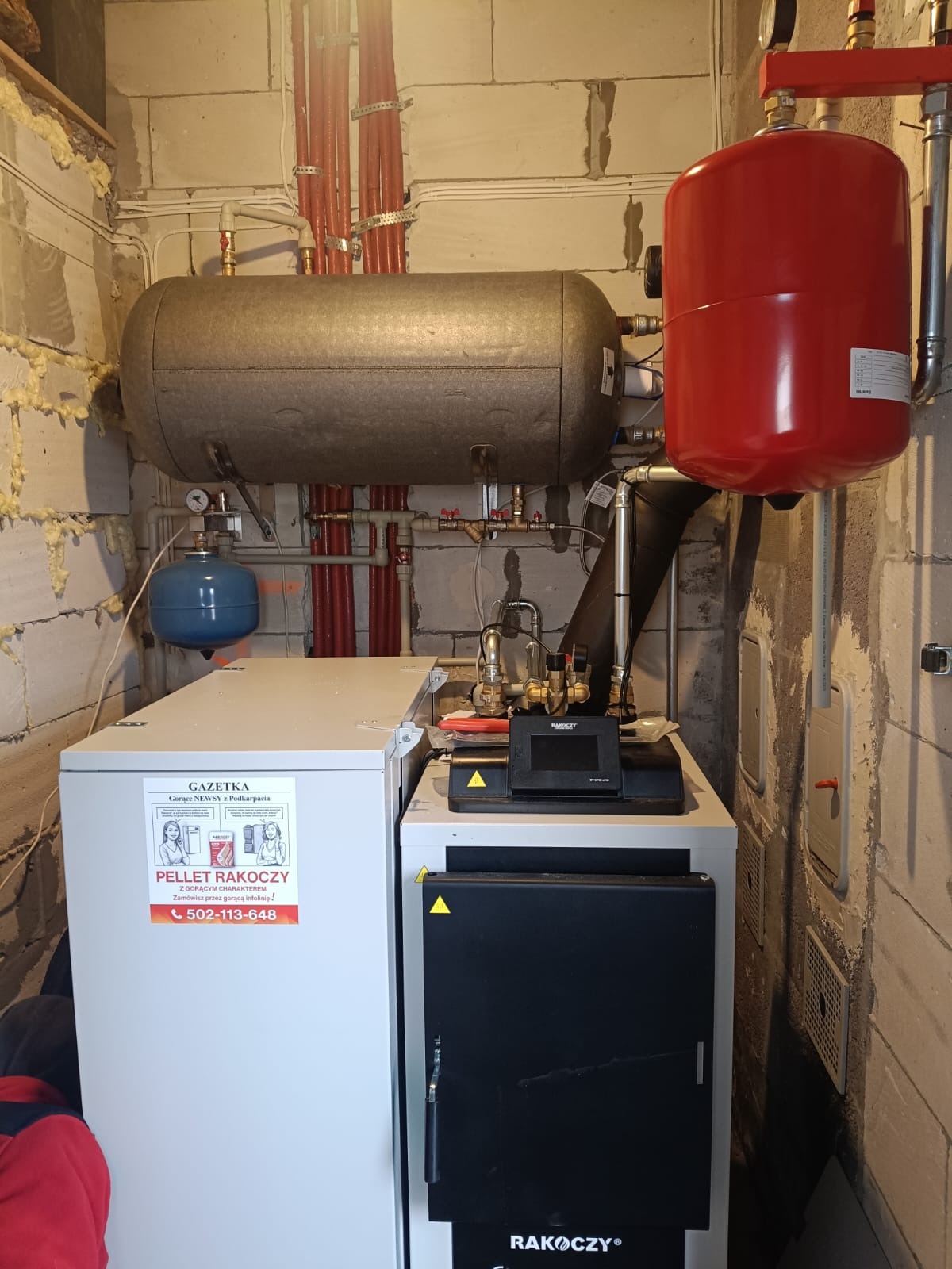 Złożona instalacja grzewcza w kotłowni: zbiorniki (szary, czerwony, niebieski), rury miedziane, piec na pellet Rakoczy z wyświetlaczem. Surowe ściany z betonu komórkowego.