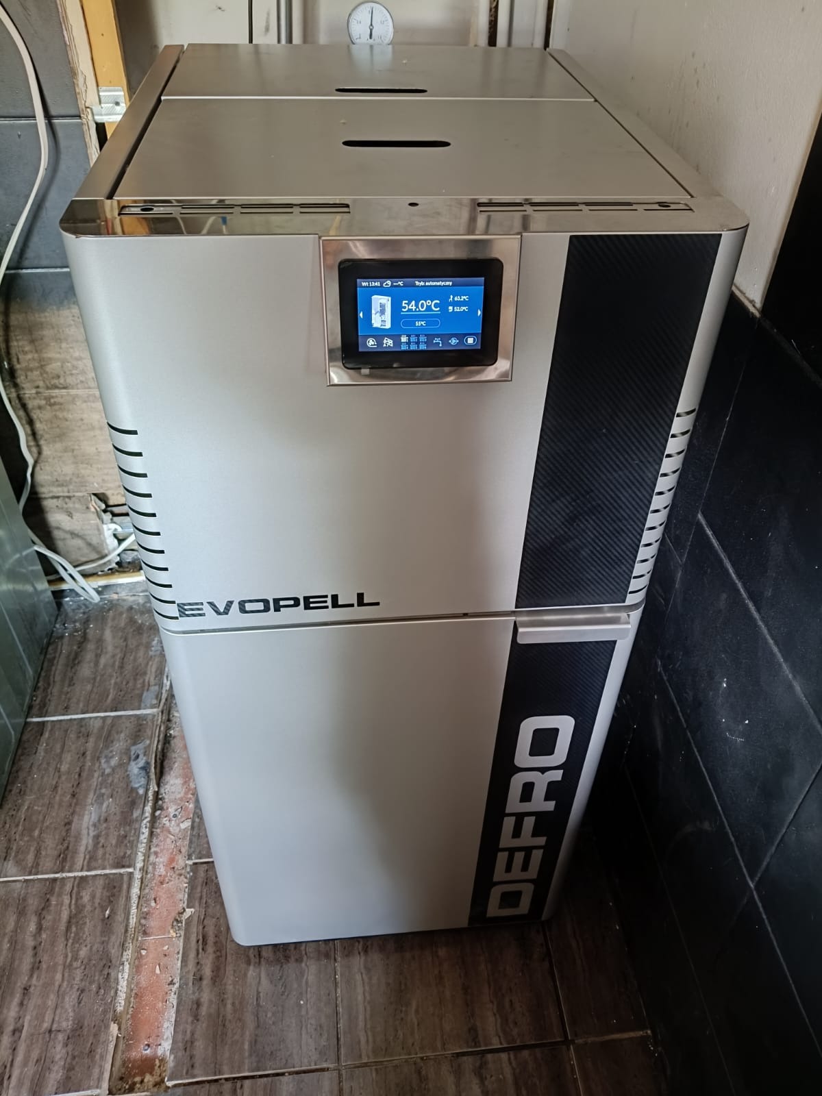 Nowoczesny piec DEFRO Evopell z wyświetlaczem temperatury 54°C, umieszczony w pomieszczeniu z brązowymi i czarnymi płytkami. Widoczny manometr na górze urządzenia.