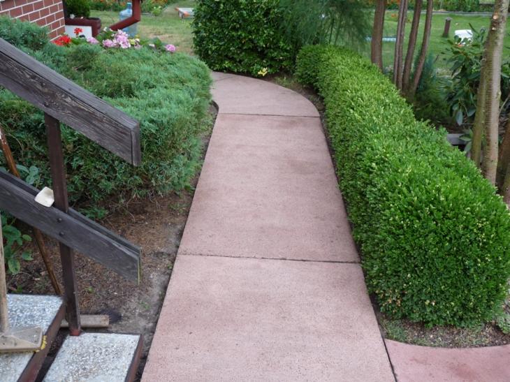 Czysta, różowa ścieżka z kostki brukowej w ogrodzie, otoczona zielenią krzewów i kwiatów. Widoczne drewniane schody z lewej strony.