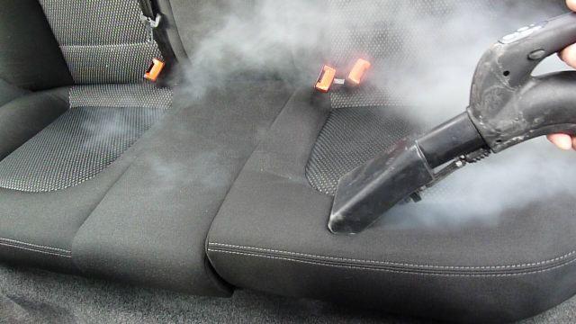Czyszczenie parowe tapicerki samochodowej. Dysza parowa w akcji, usuwająca zabrudzenia z ciemnoszarego siedzenia z widocznymi pasami i elementami zapięć.