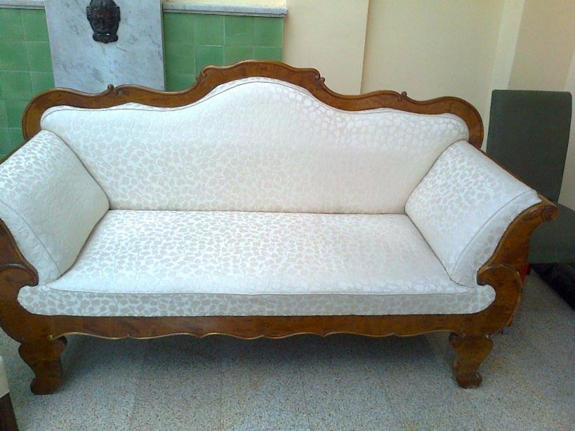 Antyczna kanapa po renowacji tapicerki w Bydgoszczy. Jasny materiał w delikatny wzór, ozdobne drewniane wykończenia. Widoczne poduszki i fragment ściany.