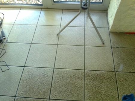 Kafelki podłogowe po czyszczeniu, widoczna różnica w czystości fug i powierzchni, fragment sprzętu czyszczącego i okna w tle, narożnik ściany po prawej.