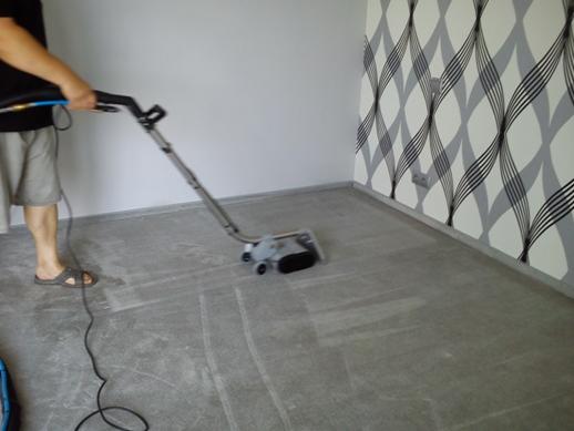 Pranie dywanu w pokoju z wzorzystą tapetą. Mężczyzna obsługuje maszynę do czyszczenia, widoczne ślady po czyszczeniu na szarym dywanie.