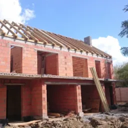 Surowy stan budynku z czerwonej cegły z drewnianą konstrukcją dachu w trakcie budowy, widoczne deski opierające się o ścianę, ziemia wokół i częściowo ogrodzenie w tle.